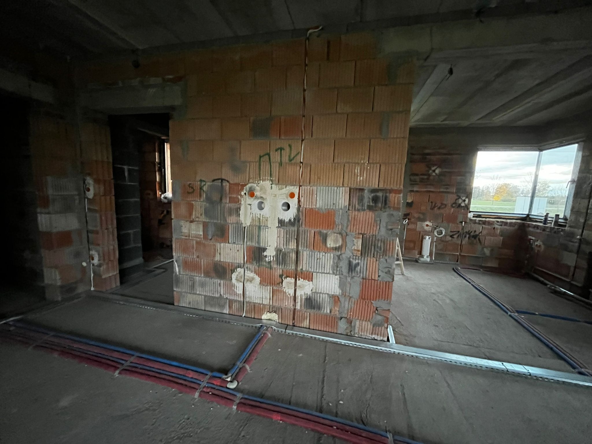 Surowy stan instalacji elektrycznej w budynku: przewody w ścianie z cegły dziurawki, rury w podłodze, widoczne puszki instalacyjne. Wnętrze w trakcie budowy.