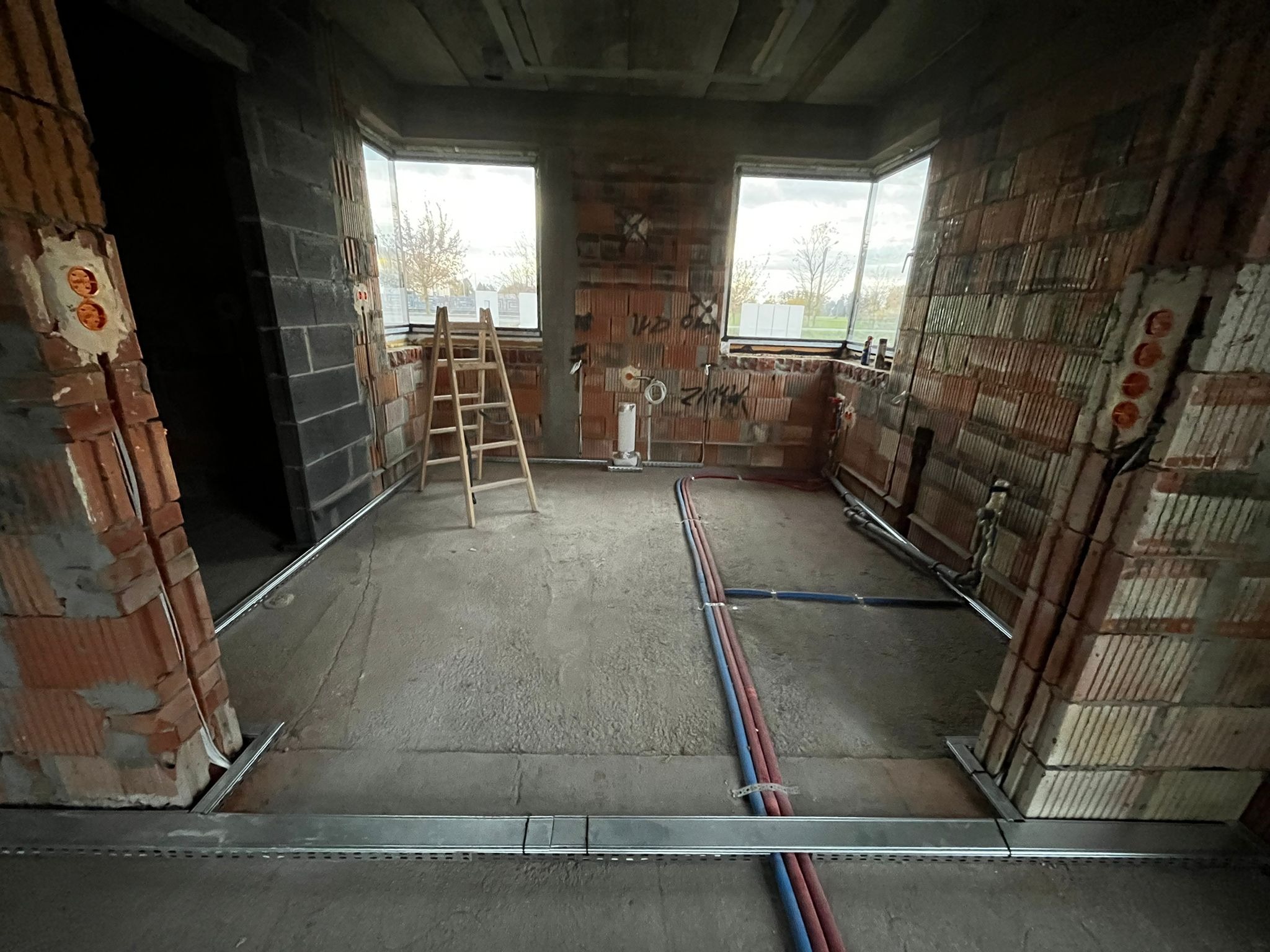 Surowy stan wnętrza z instalacjami podtynkowymi i rurami w podłodze. Widoczne puszki elektryczne w ścianach z cegły i drabina. W tle okna.