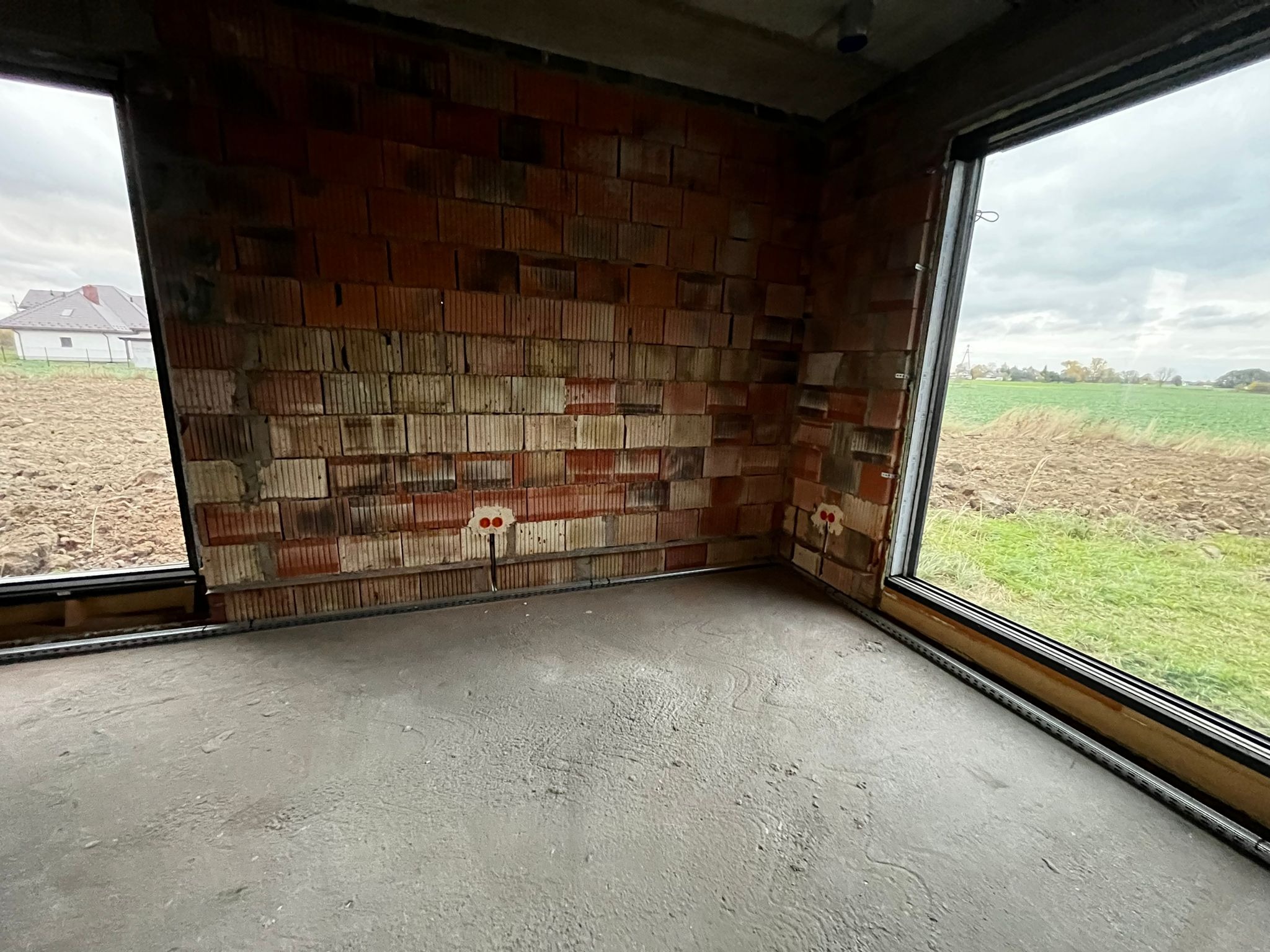 Surowy stan wnętrza: ściany z czerwonej cegły z widocznymi puszkami elektrycznymi, surowa wylewka podłogowa, okna z widokiem na pole i dom w oddali.