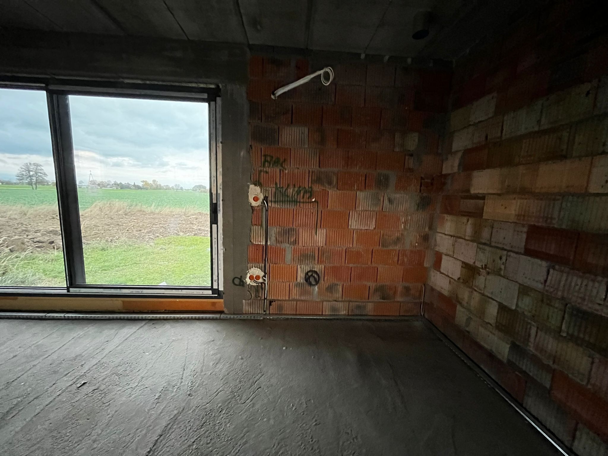 Surowy stan wnętrza z widocznymi przewodami elektrycznymi w ścianie z cegły dziurawki, okno z widokiem na pole i wylewka podłogowa. Wnętrze w trakcie prac wykończeniowych.