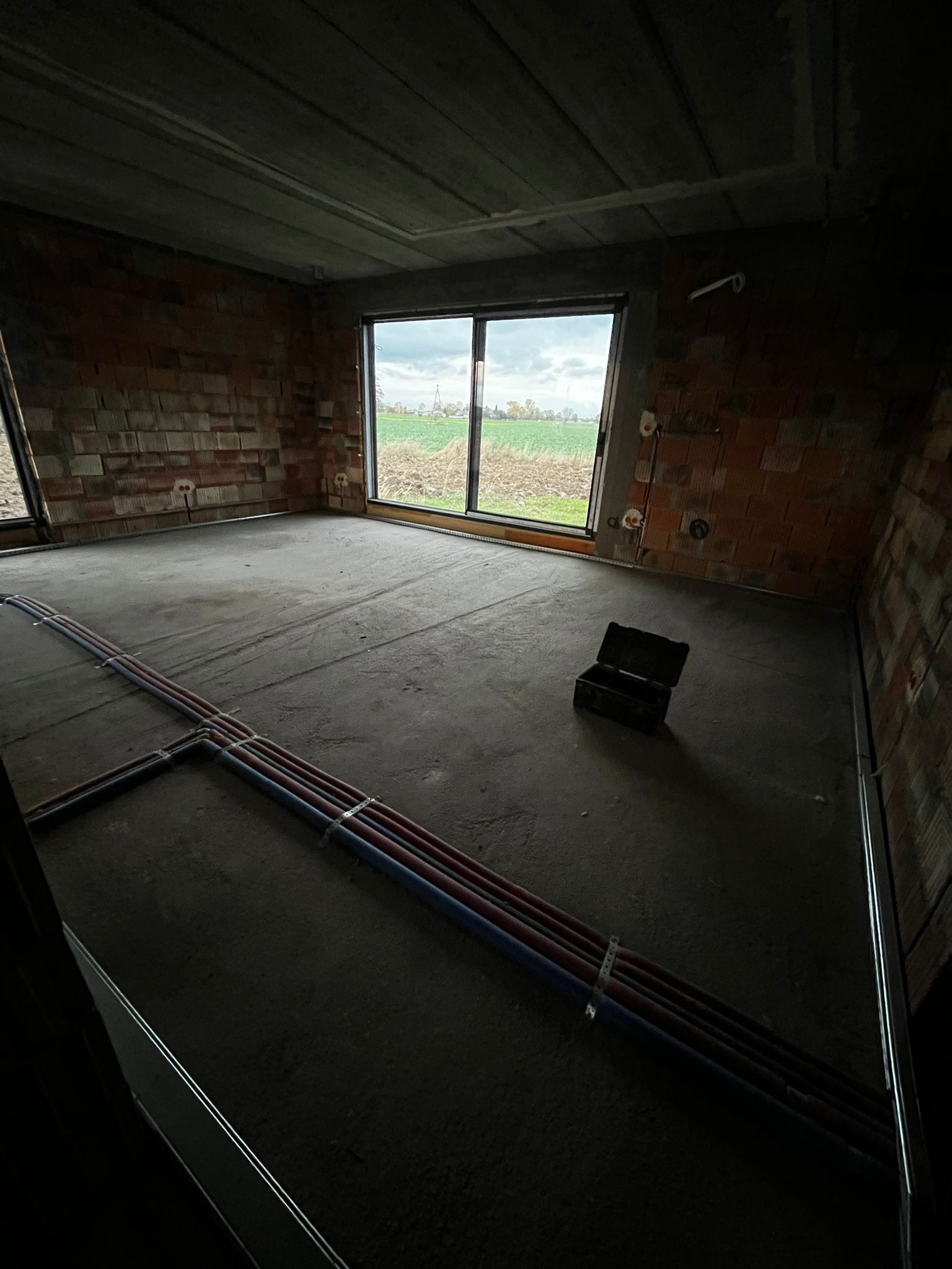 Surowy stan wnętrza z widocznymi instalacjami podłogowymi i puszkami elektrycznymi w ścianach z cegły. W tle okno z widokiem na pole. Na podłodze skrzynka narzędziowa.