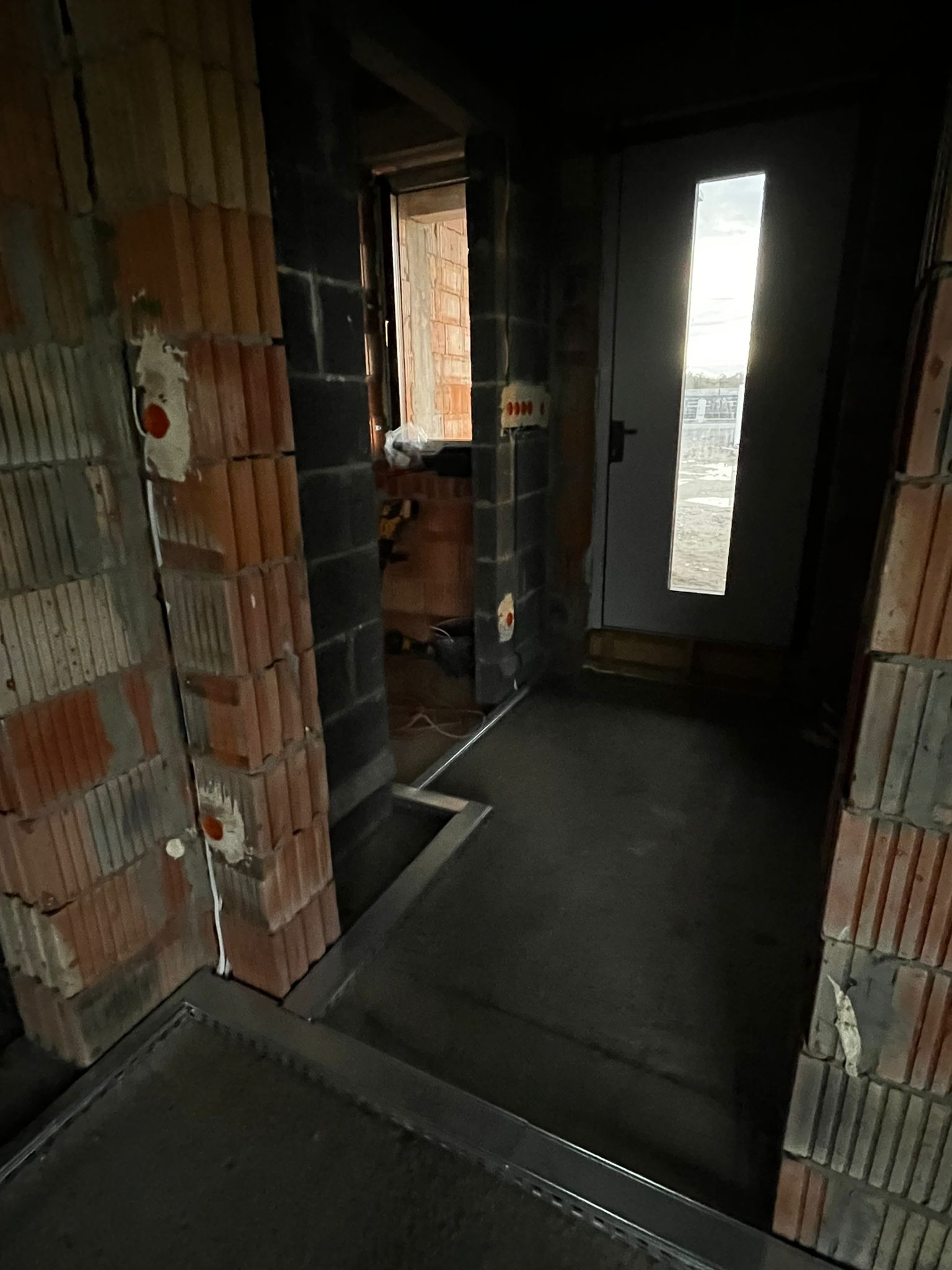 Surowy stan wnętrza budynku w trakcie budowy: widoczne ceglane ściany, instalacje elektryczne w peszlach i metalowe profile pod wylewkę. W tle okno i drzwi.