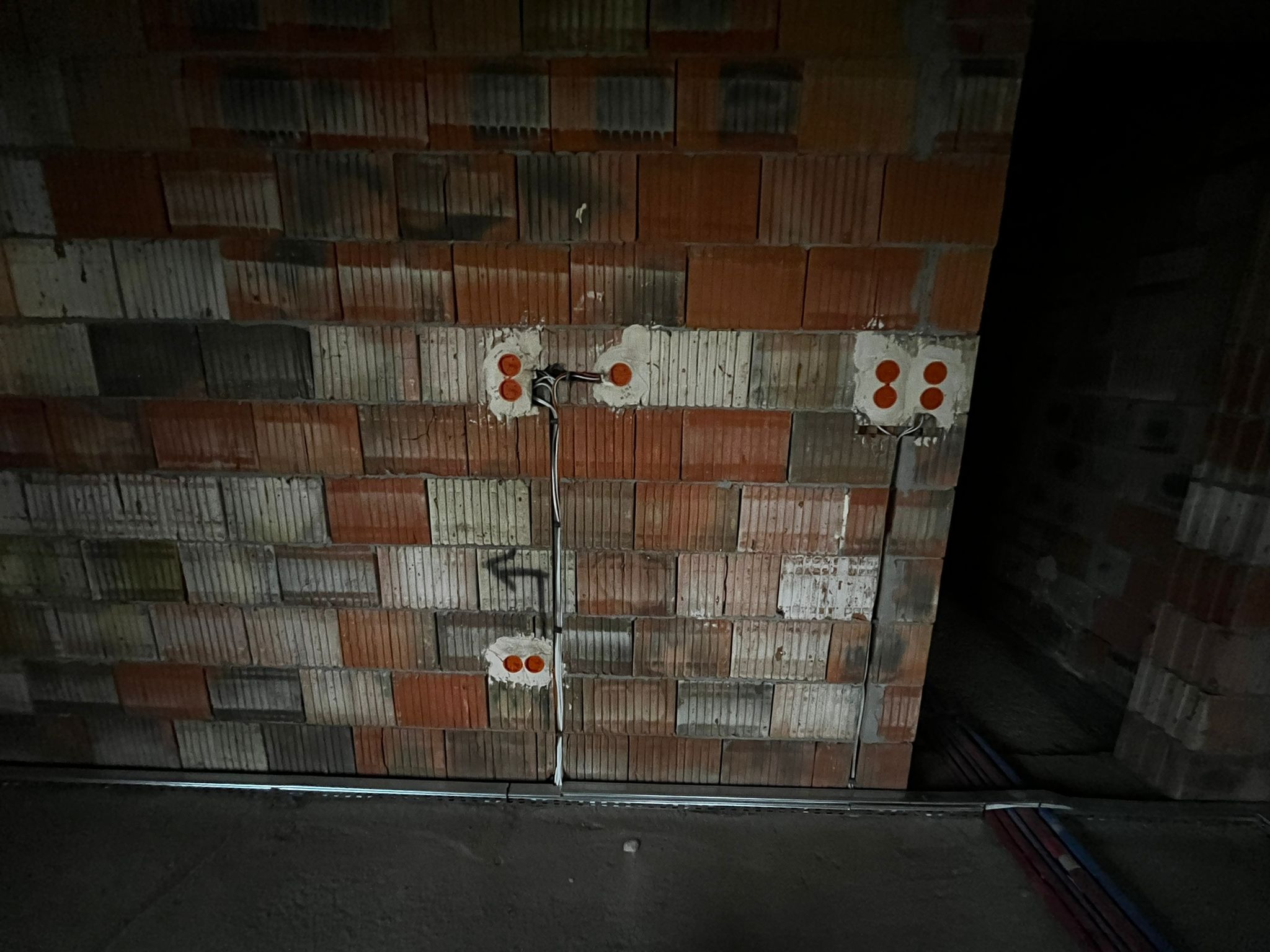 Ceglana ściana z zamontowanymi puszkami elektrycznymi i przewodami. Widoczne kable prowadzone wzdłuż ściany. Surowy stan, instalacja w trakcie realizacji.