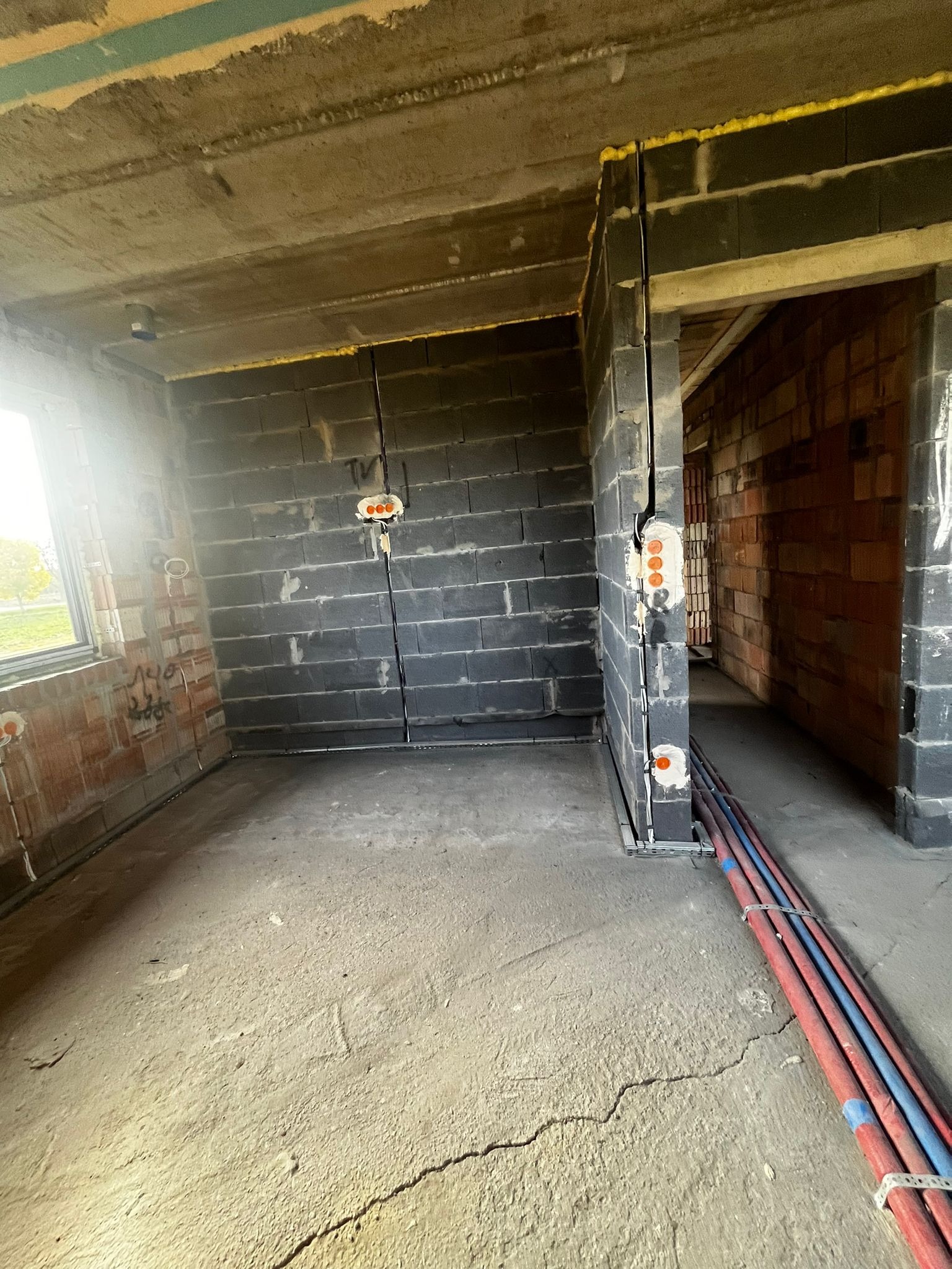 Surowy stan wnętrza z instalacją elektryczną w trakcie montażu w Sieradzu. Widoczne kable i puszki w ścianach z bloczków. Podłoże betonowe, okno z widokiem na zewnątrz.