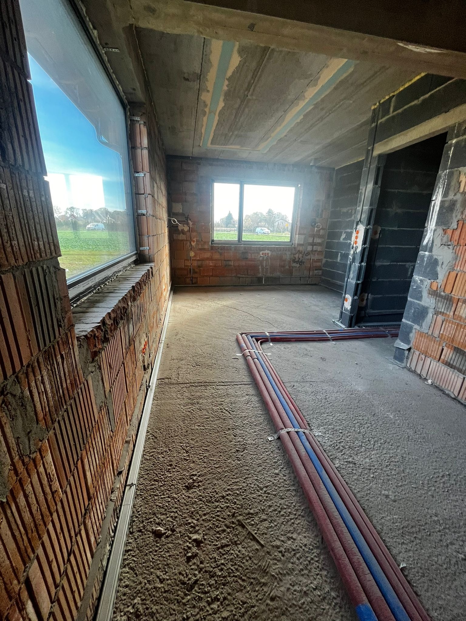Surowy stan wnętrza z widocznymi rurami instalacji grzewczej na podłodze, ściany z cegły dziurawki, okno z widokiem na zewnątrz, surowy betonowy strop.