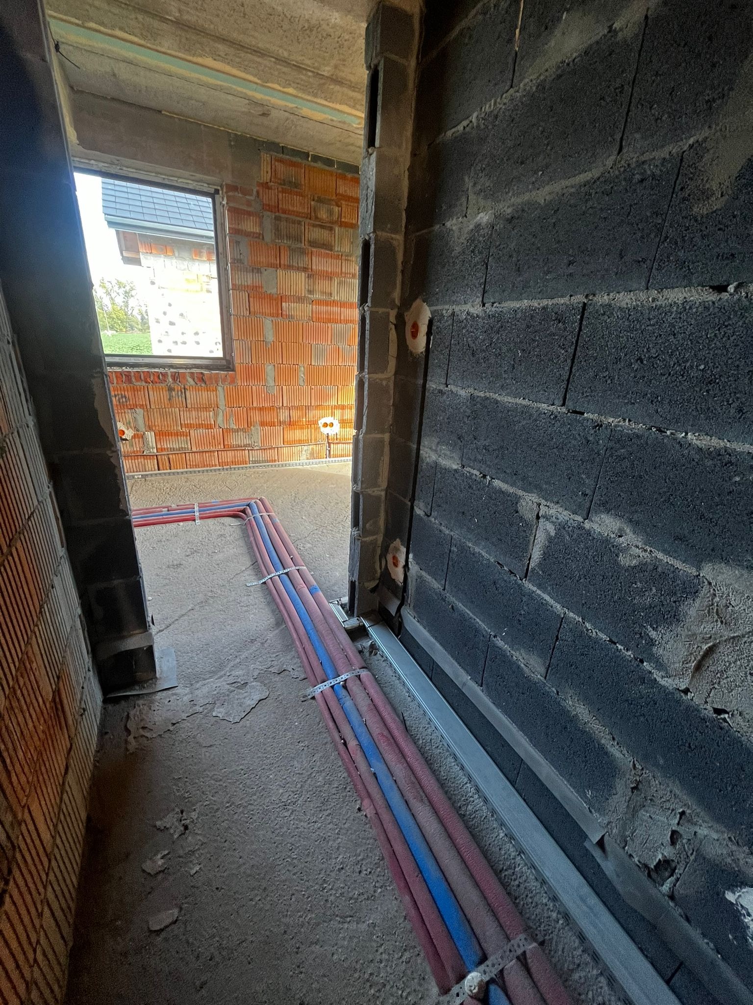 Wnętrze w trakcie budowy z widocznymi rurami instalacji grzewczej/wodnej, mocowanymi do podłoża. Ściany z cegły i bloczków betonowych, okno w tle.