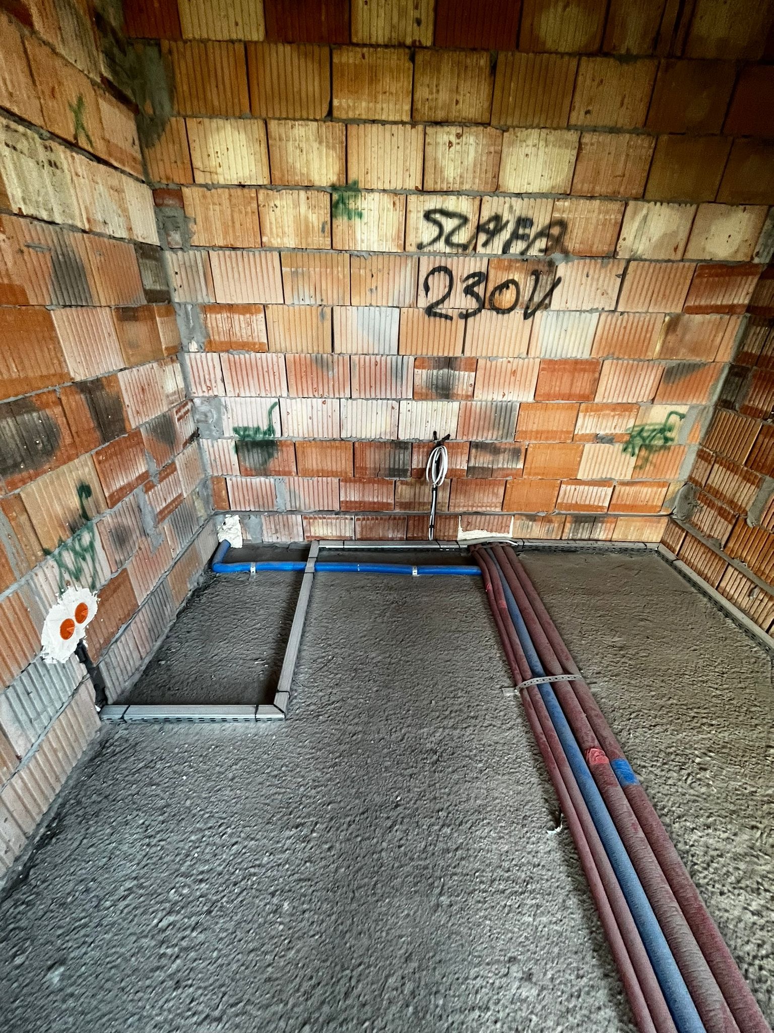 Surowy stan: instalacja rur (niebieskie, czerwone) na szarym podłożu w pomieszczeniu z ceglanymi ścianami. Widoczne gniazdka elektryczne i napisy na ścianie.