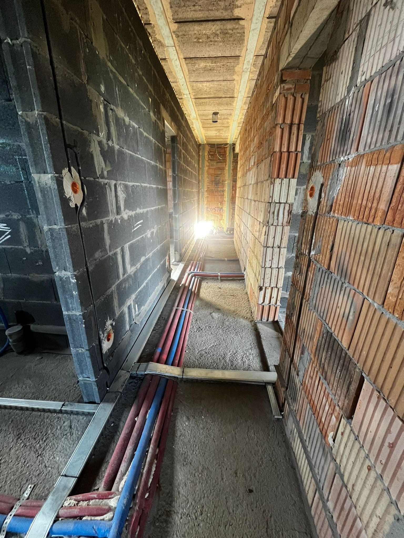 Wnętrze w trakcie budowy z widocznymi instalacjami: rurami w kolorach czerwonym i niebieskim, prowadzonymi wzdłuż ścian z cegły i pustaków. Widoczne otwory pod gniazdka.