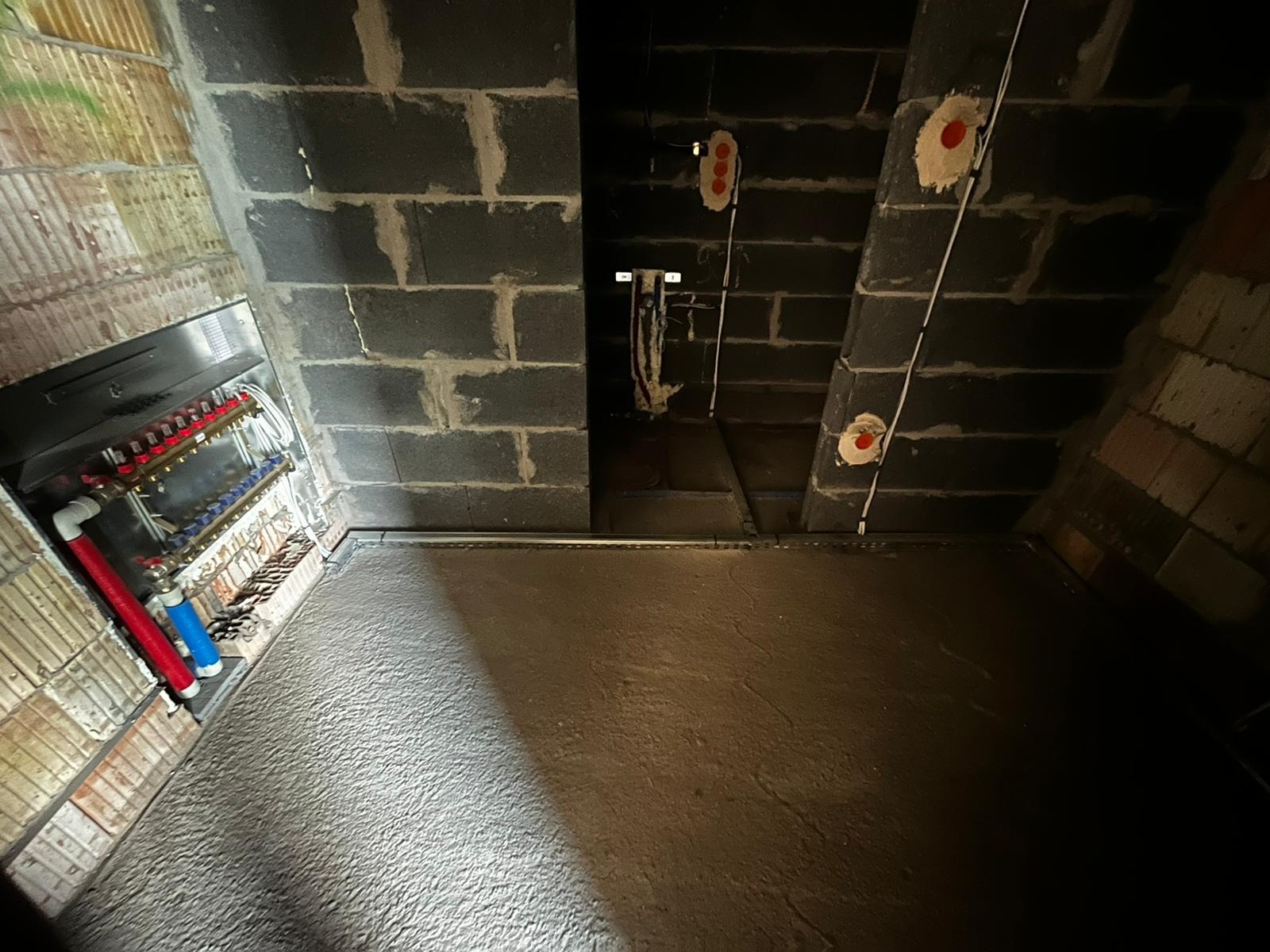 Surowy stan poddasza z instalacją elektryczną i ogrzewaniem podłogowym: widoczne przewody, puszki elektryczne na ścianie z bloczków oraz rozdzielacz ogrzewania podłogowego.