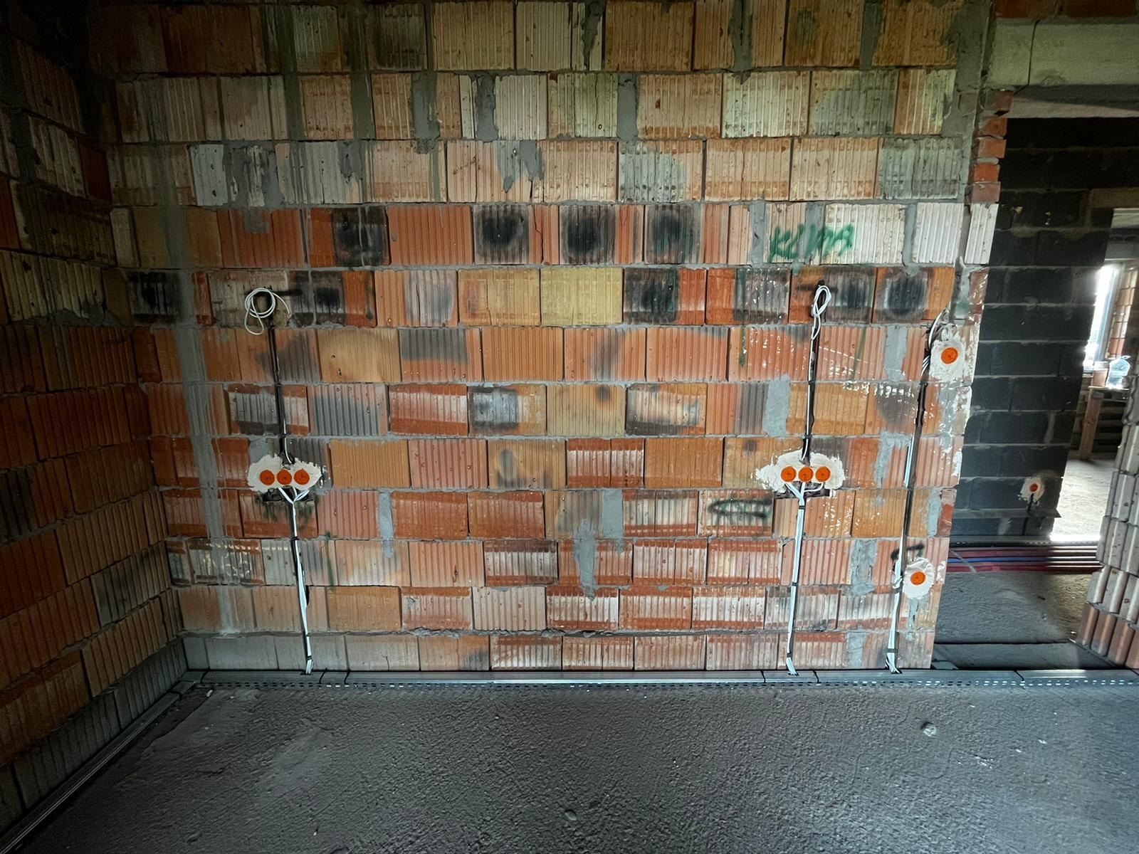 Wewnętrzna ściana z cegły z widocznymi puszkami instalacyjnymi i przewodami elektrycznymi, przygotowana do dalszych prac wykończeniowych. Surowy stan.