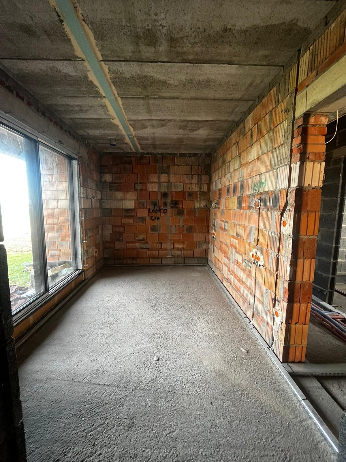 Surowy stan wnętrza: ściany z czerwonej cegły z widocznymi puszkami elektrycznymi, betonowa podłoga i sufit, duże okno z widokiem na zewnątrz. Wnętrze w trakcie prac wykończeniowych.