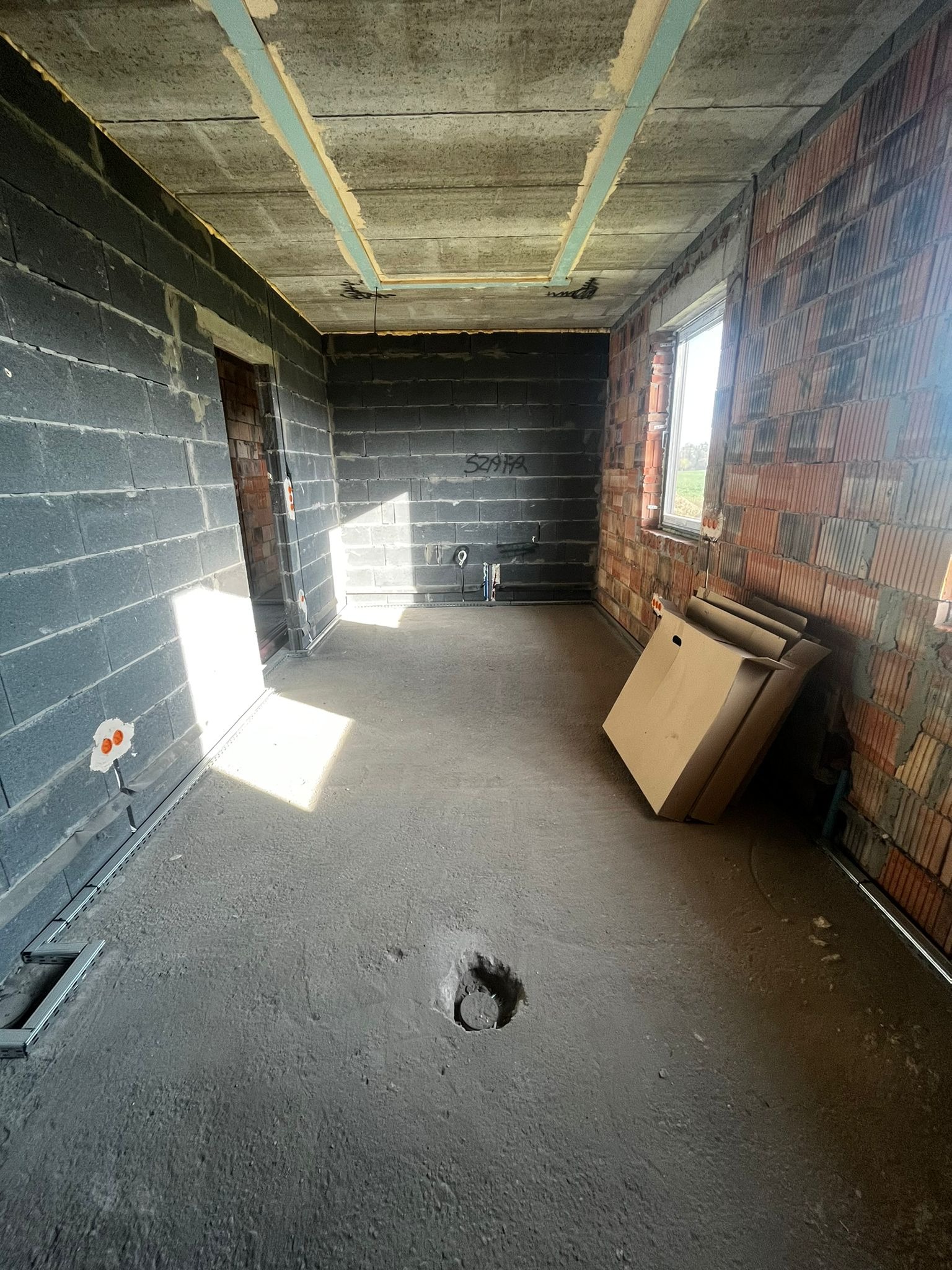 Surowy stan wnętrza budynku w trakcie budowy: ściany z bloczków i cegieł, betonowa podłoga z otworem, widoczne instalacje elektryczne, okno z widokiem na zewnątrz.