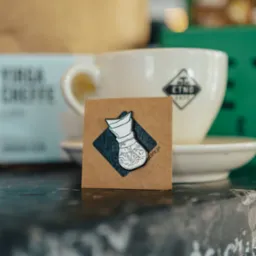 Emailiowana przypinka przedstawiająca filtr do kawy Chemex z motywem górskiego krajobrazu na kartonowym tle, obok filiżanki z logo ETNO Cafe.