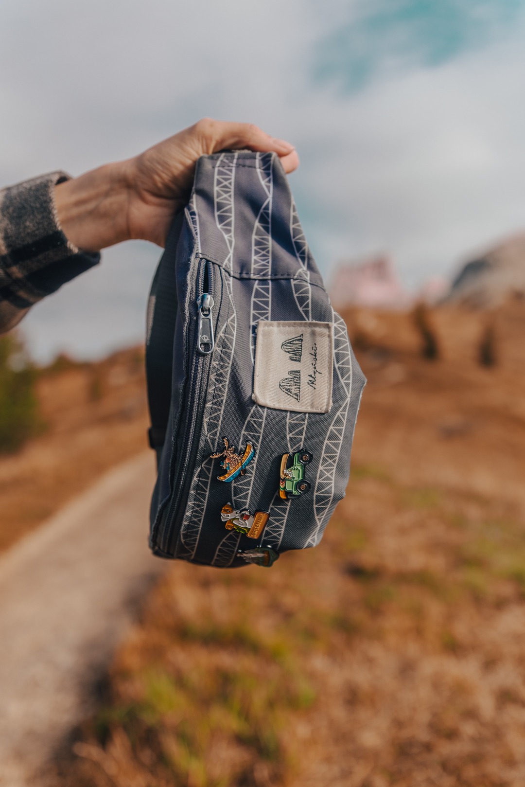 Szara torba typu nerka z białym wzorem przypominającym konstrukcję stalową, ozdobiona trzema przypinkami z motywami rekreacyjnymi, trzymana w dłoni na tle górskiego krajobrazu.