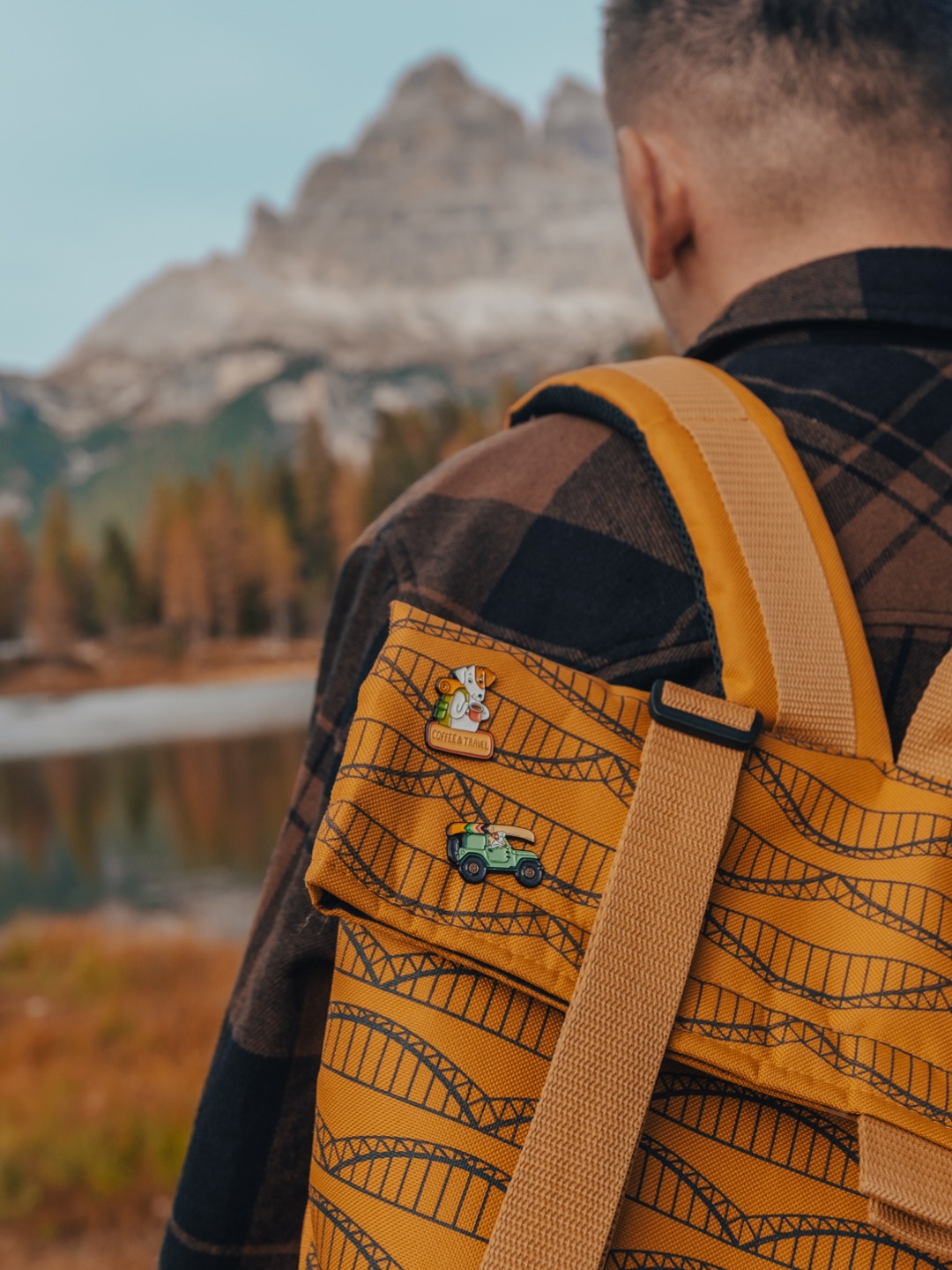 Mężczyzna z plecakiem z przypinkami na tle górskiego krajobrazu z jeziorem i jesiennymi drzewami. Na plecaku dwie przypinki: pies z plecakiem i napisem 'Coffee & Travel' oraz zielony jeep z deską...