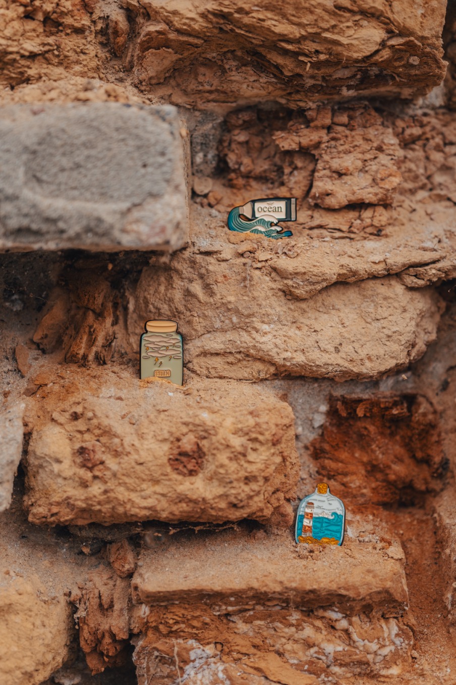 Trzy emaliowane przypinki przedstawiające motywy oceaniczne (fala z napisem ocean, burza w słoiku i latarnia morska w butelce) umieszczone na starej, ceglanej ścianie.