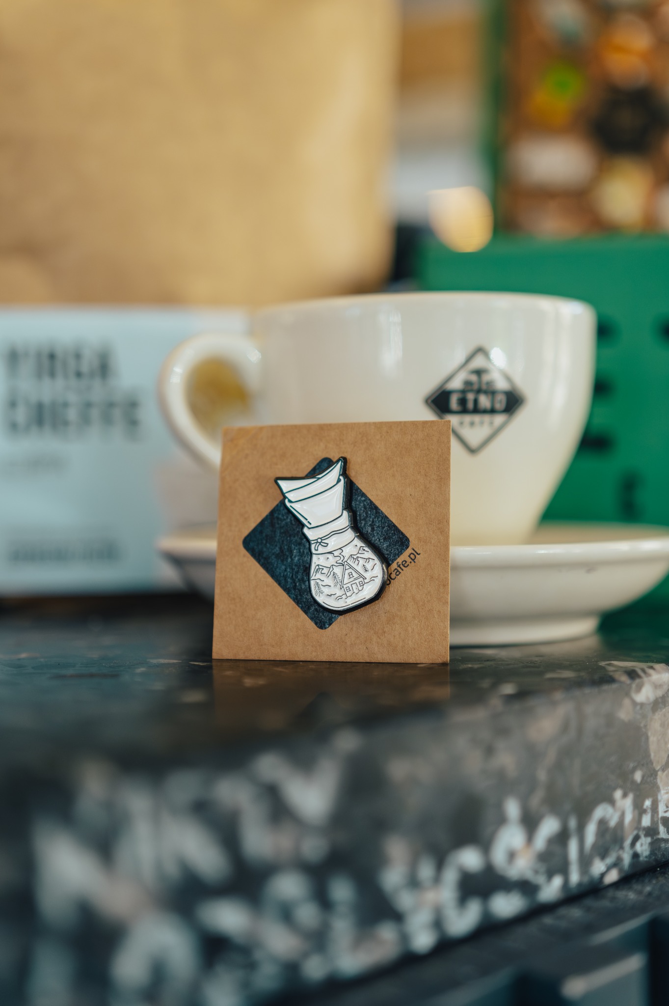 Emailiowana przypinka przedstawiająca filtr do kawy Chemex z motywem górskiego krajobrazu na kartonowym tle, obok filiżanki z logo ETNO Cafe.
