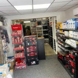 Wnętrze sklepu z artykułami hydraulicznymi, widoczne regały z rurami, złączkami, narzędziami i skrzynkami narzędziowymi firmy Fischer, oświetlone lampami sufitowymi.