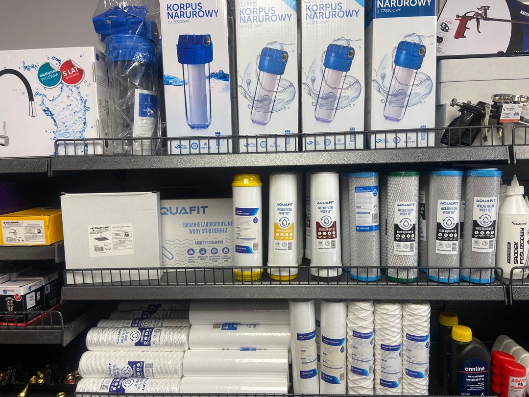 Półki w sklepie z artykułami hydraulicznymi, prezentujące filtry do wody, wkłady filtrujące i obudowy filtrów narurowych, wraz z opakowaniami laboratoryjnych badań wody.