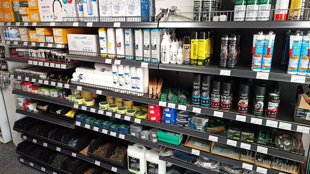 Półki w sklepie z artykułami hydraulicznymi, prezentujące filtry do wody, uszczelniacze, silikony, grunty, narzędzia i akcesoria instalacyjne, uporządkowane według kategorii i producentów.