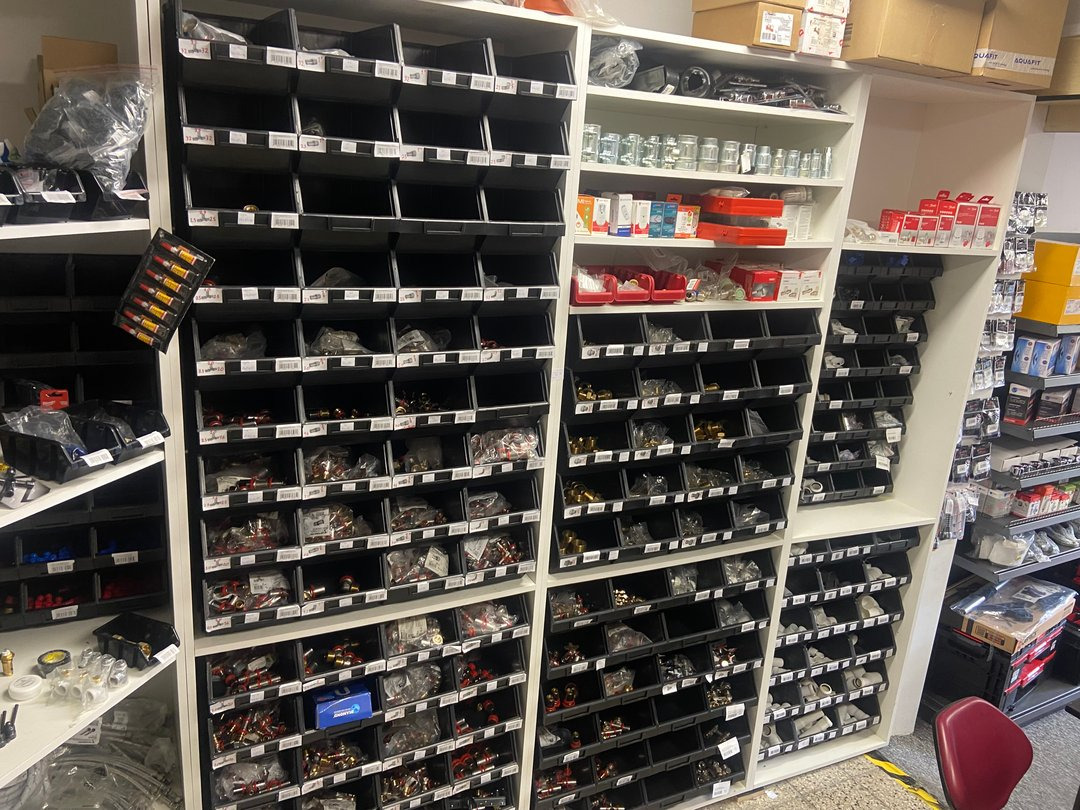 Półki regału w sklepie, wypełnione różnorodnymi złączkami, rurkami i akcesoriami hydraulicznymi, posegregowanymi w czarnych pojemnikach z etykietami, widoczne także pudełka z produktami na górnych...