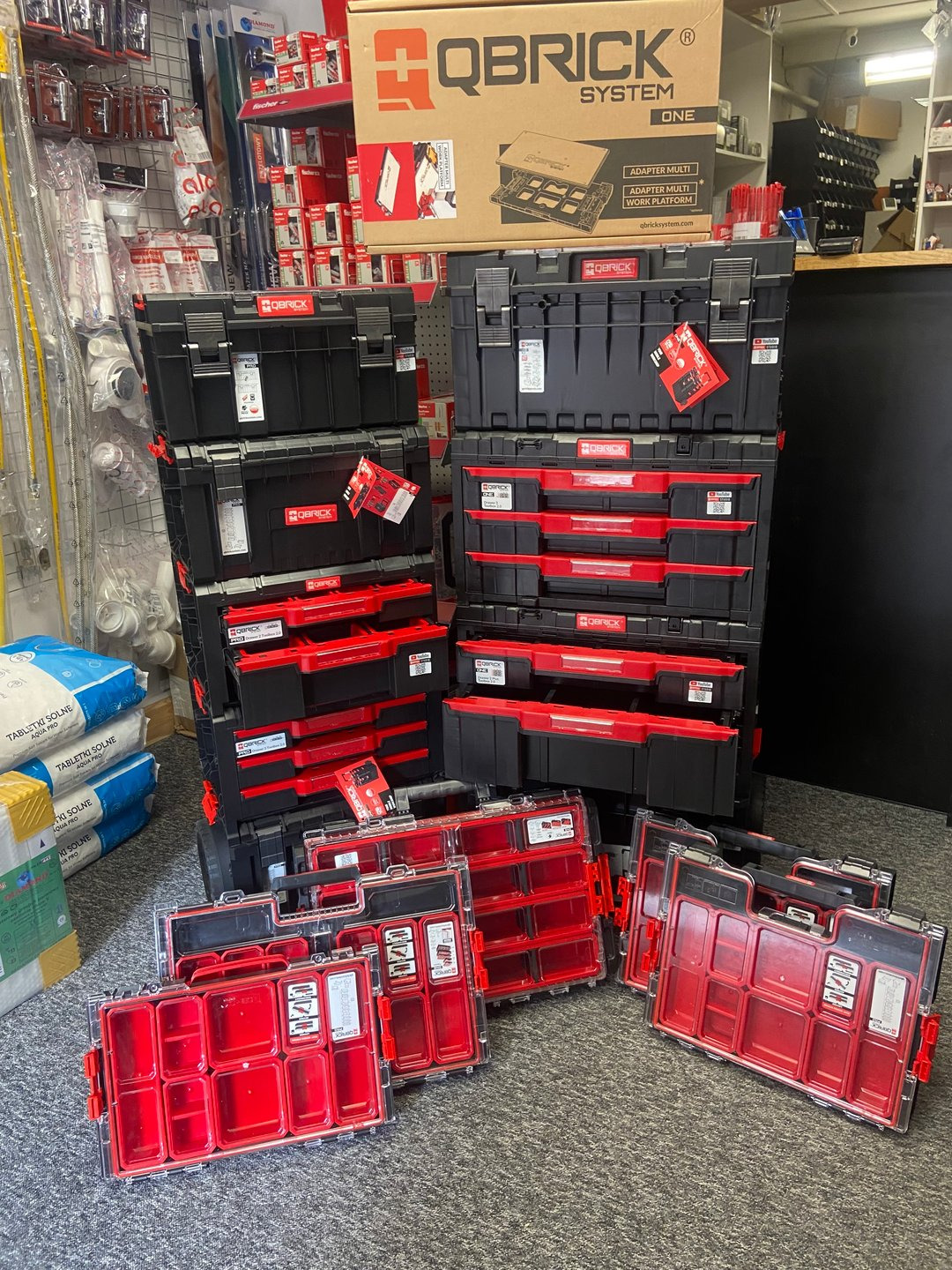 Ekspozycja zestawu modułowych skrzynek narzędziowych Qbrick System ONE, z czarnymi korpusami i czerwonymi elementami, ustawionych w stosy i rozłożonych na podłodze w sklepie.