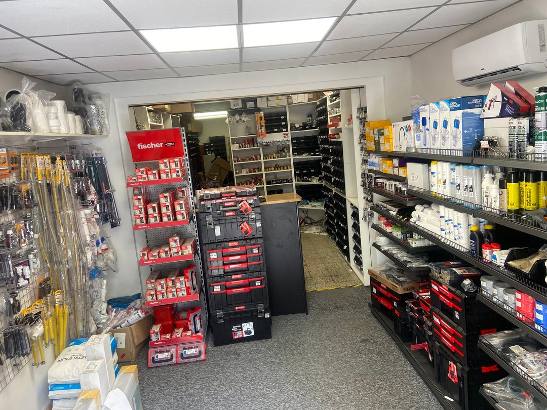 Wnętrze sklepu z artykułami hydraulicznymi, widoczne regały z rurami, złączkami, narzędziami i skrzynkami narzędziowymi firmy Fischer, oświetlone lampami sufitowymi.