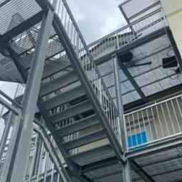 Zewnętrzne, metalowe schody ewakuacyjne z ażurowymi stopniami i balustradą, przymocowane do budynku z oknem w kolorze błękitu.