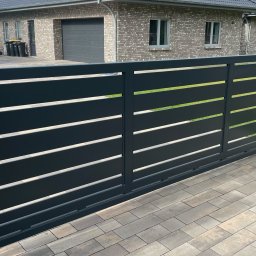 METALLBAU LESZEK CHROSTOWSKI - Nowoczesna, przesuwna brama w kolorze antracytu z poziomymi szczelinami, zamontowana przy ceglanym słupku wjazdowym na posesję z brukowanym podjazdem i widocznym budynkiem mieszkalnym w tle.