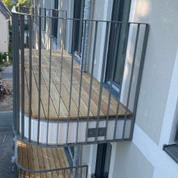 METALLBAU LESZEK CHROSTOWSKI - Nowoczesny balkon z drewnianą podłogą i szarą metalową balustradą, widok z góry na dwa balkony, fragment elewacji budynku z oknami w tle.