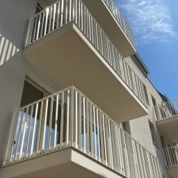 METALLBAU LESZEK CHROSTOWSKI - Nowoczesne balkony w budynku mieszkalnym z białymi, metalowymi balustradami o pionowym układzie szczebli, widziane z dołu pod kątem w słoneczny dzień.
