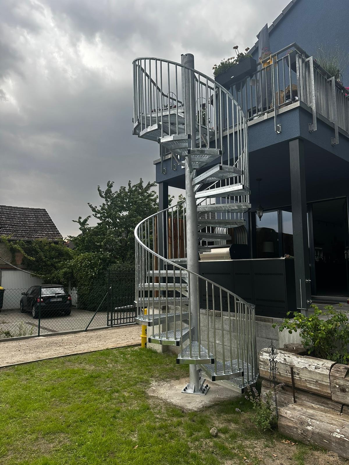 Zewnętrzne metalowe schody spiralne prowadzące na balkon domu z szarą elewacją, widoczne elementy mocujące do podłoża.