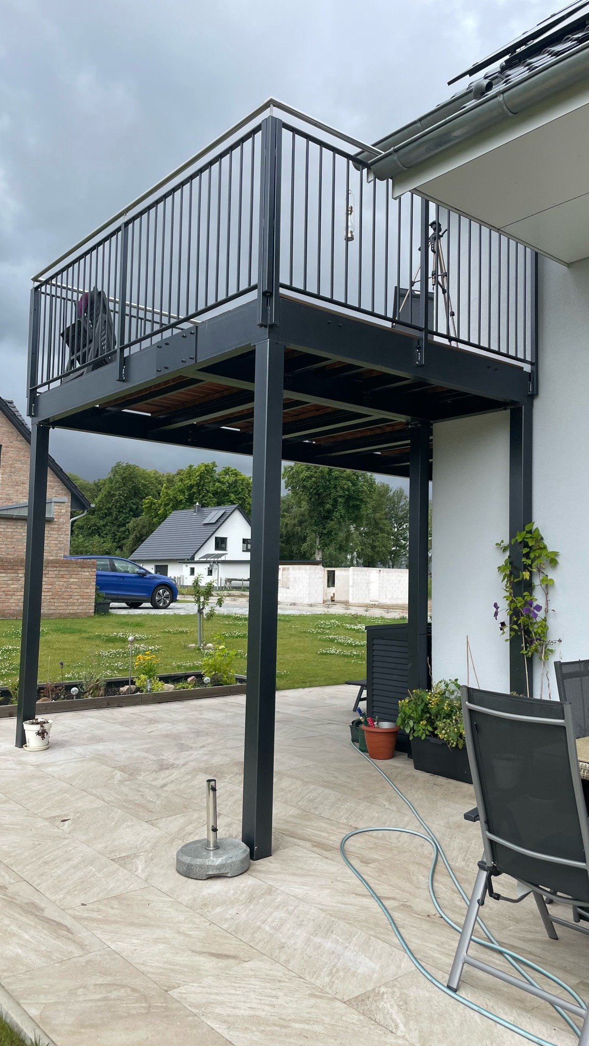 Nowoczesny balkon z czarną, metalową balustradą na tle pochmurnego nieba i zadbanego ogrodu, widoczny z poziomu tarasu.
