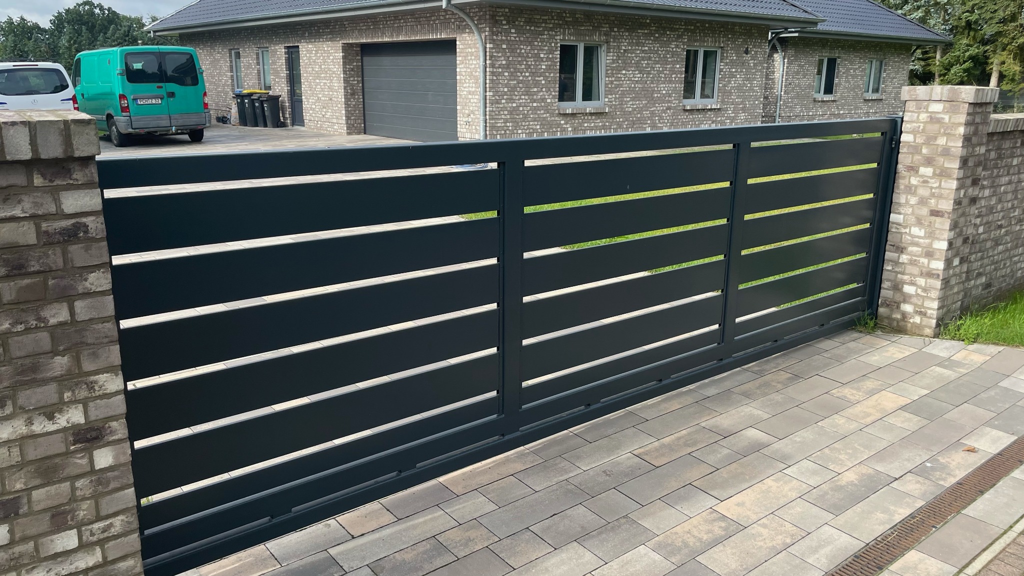 Nowoczesna, przesuwna brama w kolorze antracytu z poziomymi szczelinami, zamontowana przy ceglanym słupku wjazdowym na posesję z brukowanym podjazdem i widocznym budynkiem mieszkalnym w tle.
