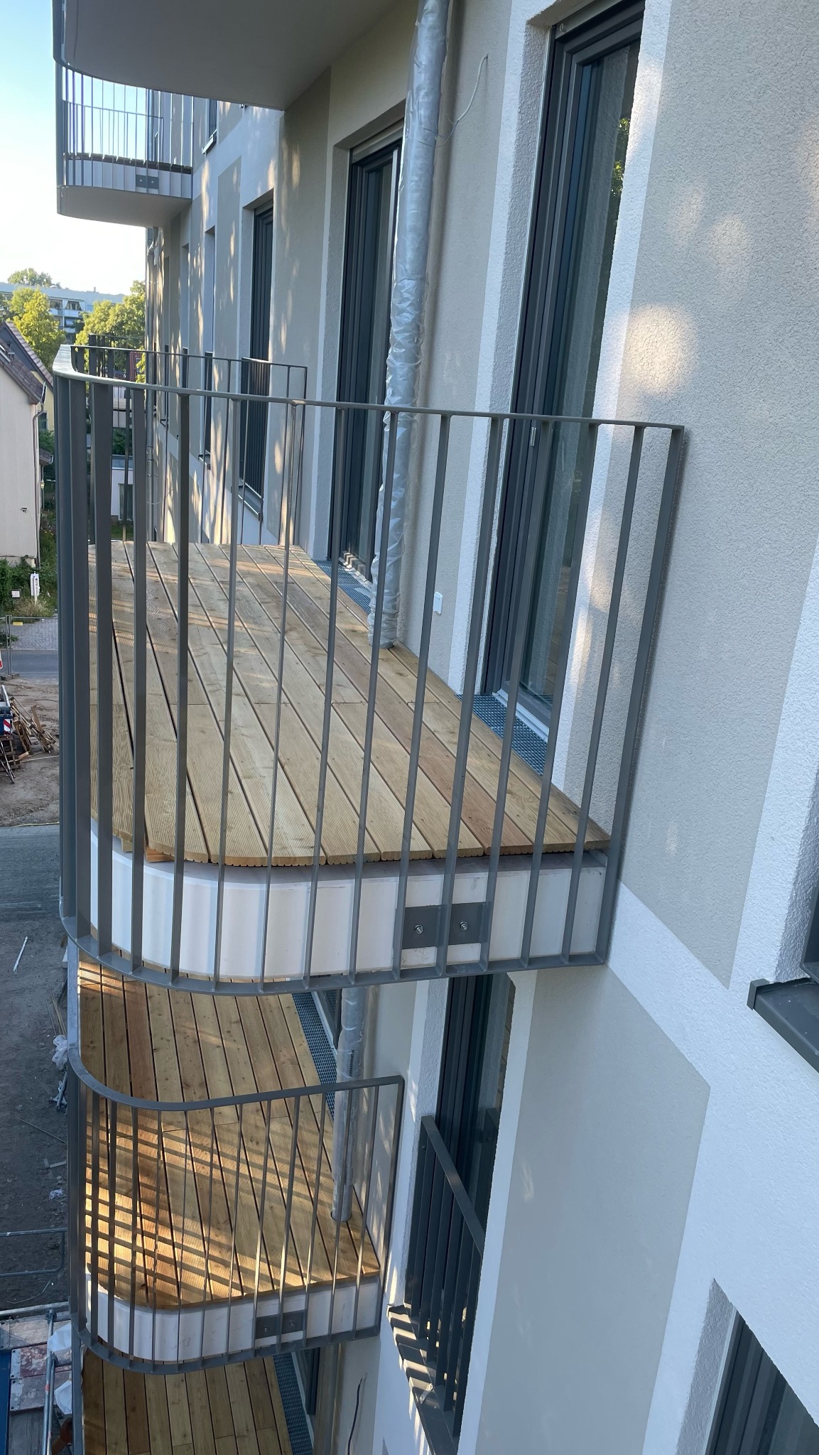 Nowoczesny balkon z drewnianą podłogą i szarą metalową balustradą, widok z góry na dwa balkony, fragment elewacji budynku z oknami w tle.