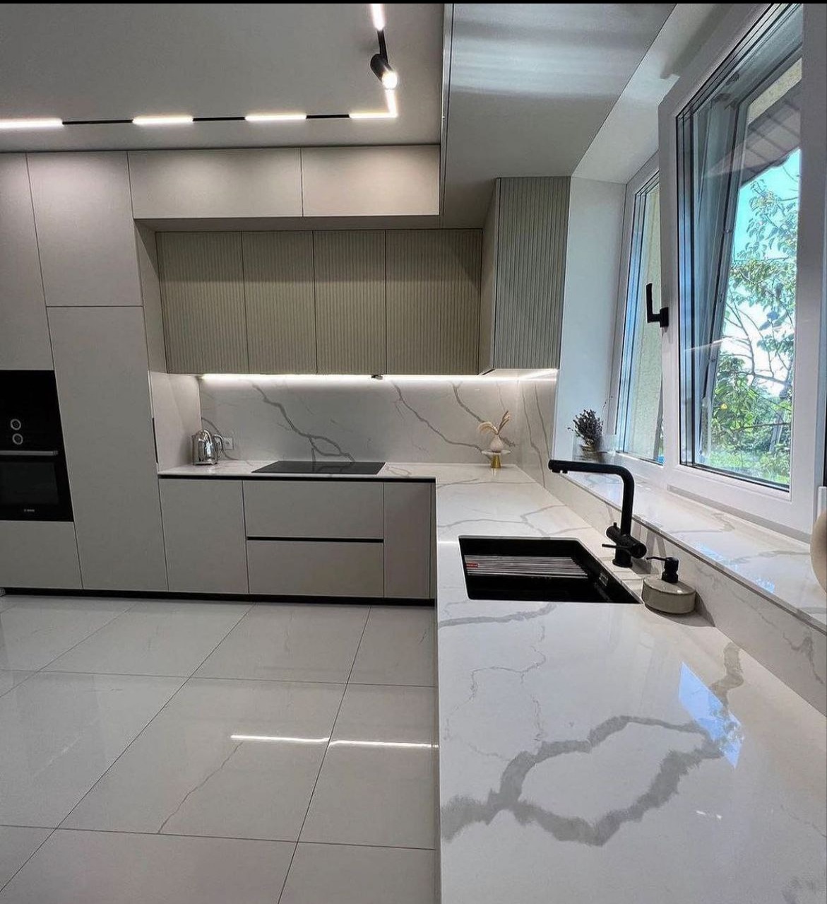 Jasna kuchnia z białym marmurowym blatem, czarnym zlewem i baterią, minimalistycznym oświetleniem LED i widokiem na zieleń za oknem.