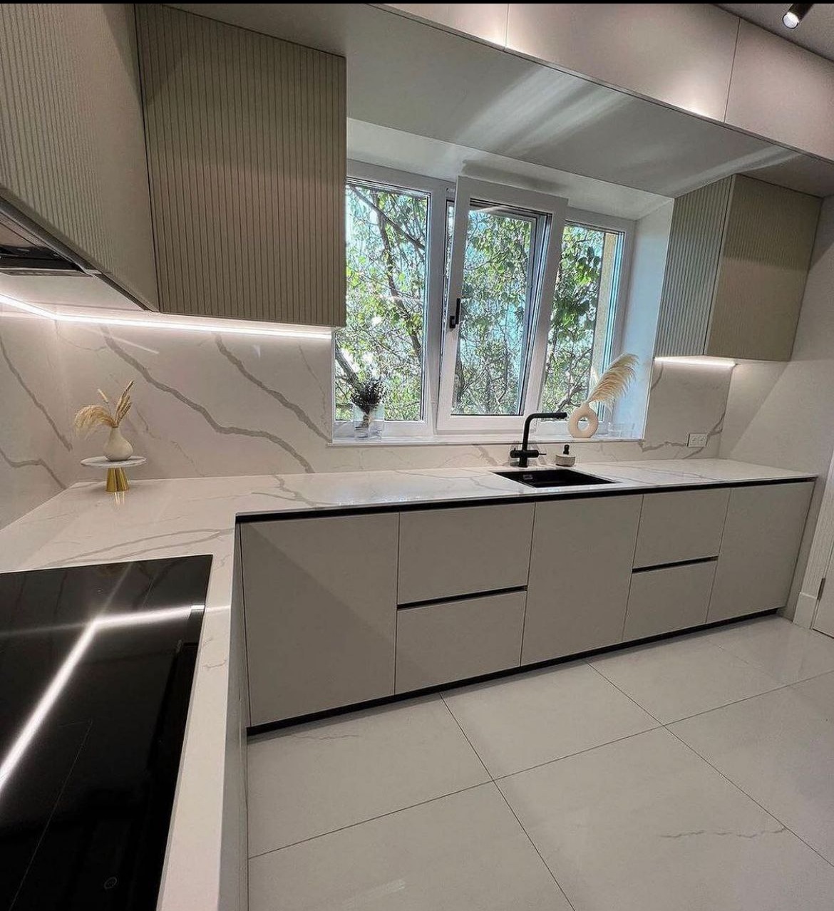 Nowoczesna kuchnia z beżowymi frontami, marmurowym blatem i backsplashiem, czarną armaturą i widokiem na zieleń za oknem.