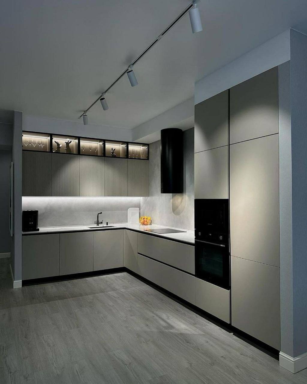 Nowoczesna kuchnia w odcieniach szarości z oświetleniem szynowym, czarnym okapem i przeszklonymi szafkami wiszącymi z dekoracjami.