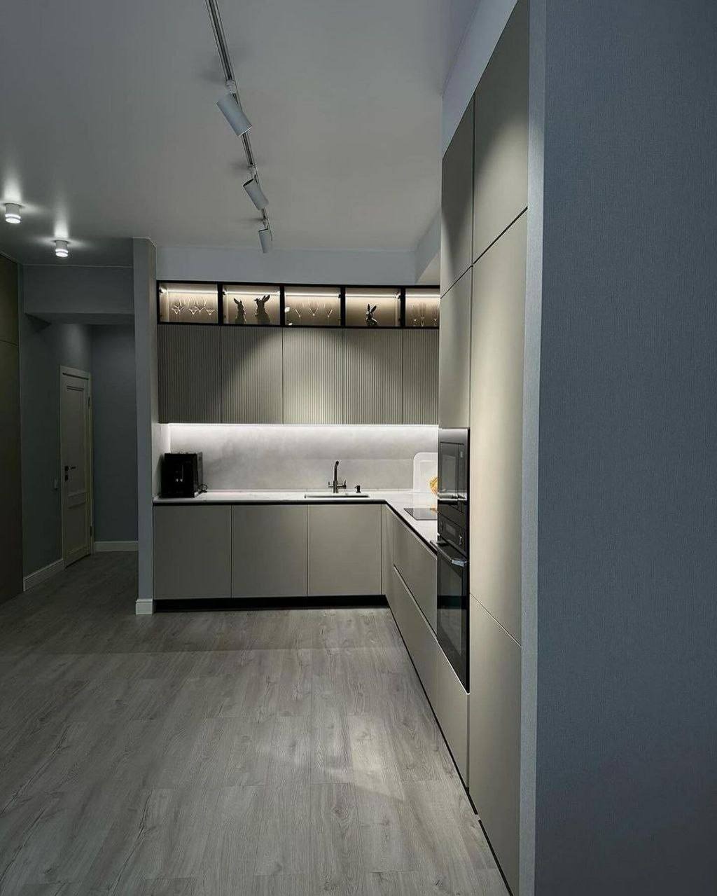 Nowoczesna kuchnia w odcieniach szarości z podświetlanymi szafkami, dekoracyjnymi figurkami w górnej części i drewnopodobną podłogą.