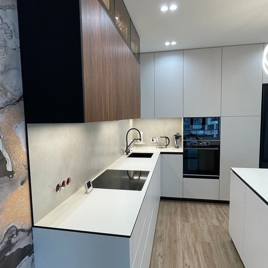 Nowoczesna zabudowa kuchenna z białymi frontami, czarnymi akcentami i drewnianymi szafkami wiszącymi, widoczne gniazdka elektryczne w ścianie.