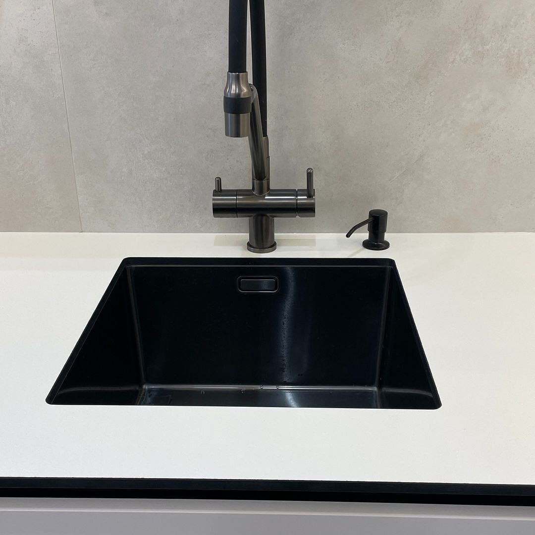 Czarny, trapezoidalny zlew kuchenny z ciemnoszarą baterią i dozownikiem mydła na białym blacie, minimalistyczny design, szary panel ścienny w tle.