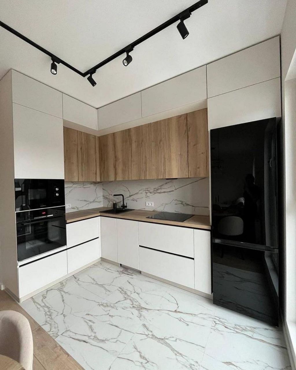 Nowoczesna kuchnia narożna z białymi i drewnianymi szafkami, czarnym sprzętem AGD i marmurową podłogą, oświetlona reflektorami sufitowymi.