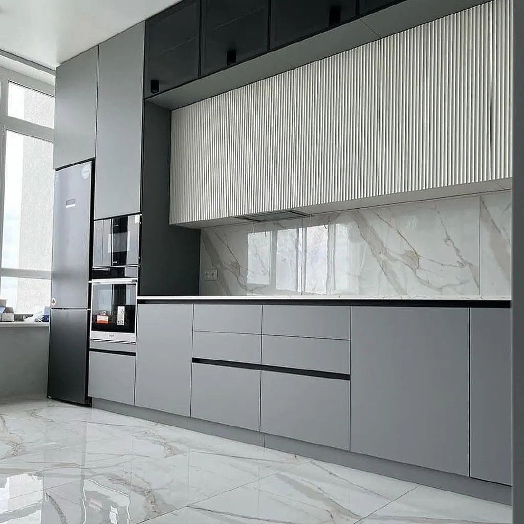 Szara, nowoczesna zabudowa kuchenna z białymi, marmurowymi płytkami na ścianie i podłodze, oświetlenie LED pod szafkami, czarne akcenty na uchwytach i górnych szafkach.