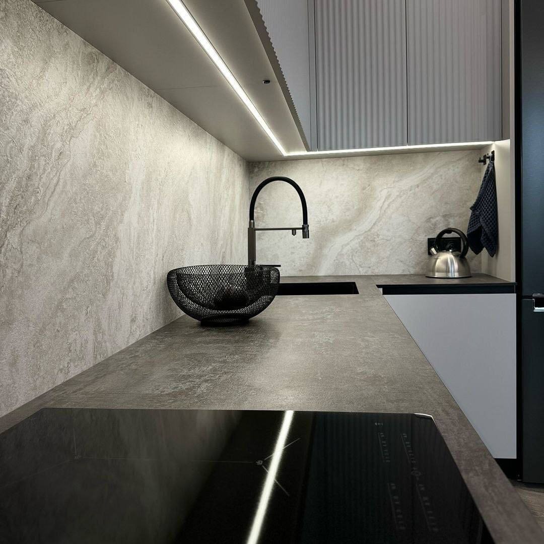 Minimalistyczna kuchnia z szarą, kamienną ścianą, czarną baterią i koszykiem na owoce, oświetlenie LED pod szafkami