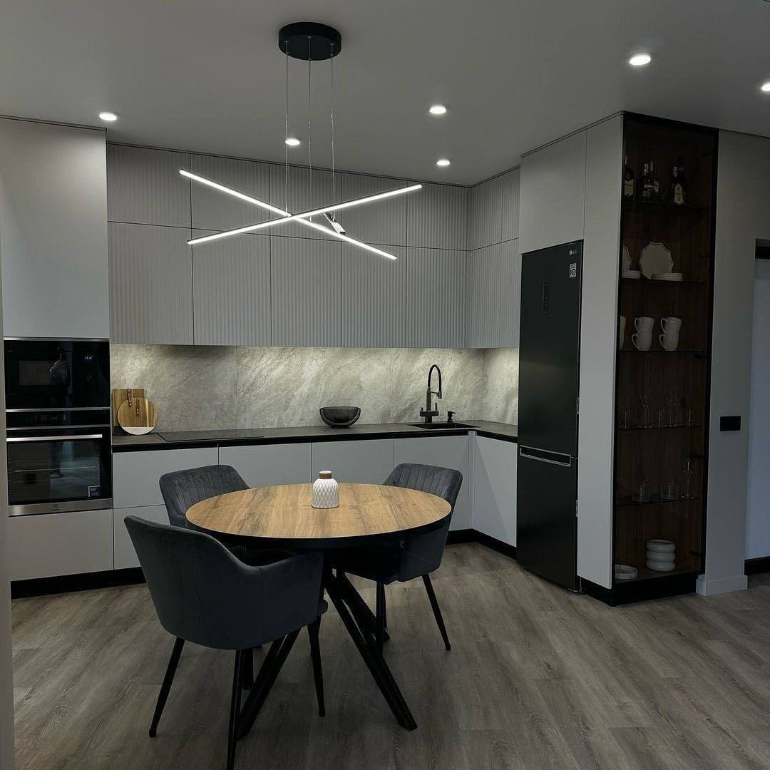 Nowoczesna kuchnia z szarymi frontami, okrągłym drewnianym stołem i designerską lampą sufitową w kształcie skrzyżowanych linii.