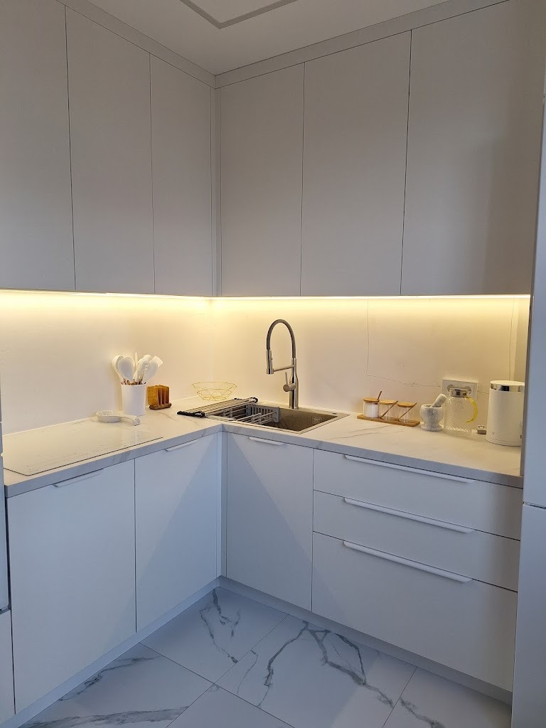 Jasna, minimalistyczna kuchnia w kształcie litery L z białymi frontami szafek, marmurową podłogą i oświetleniem podszafkowym, widok na narożnik ze zlewozmywakiem i akcesoriami kuchennymi.