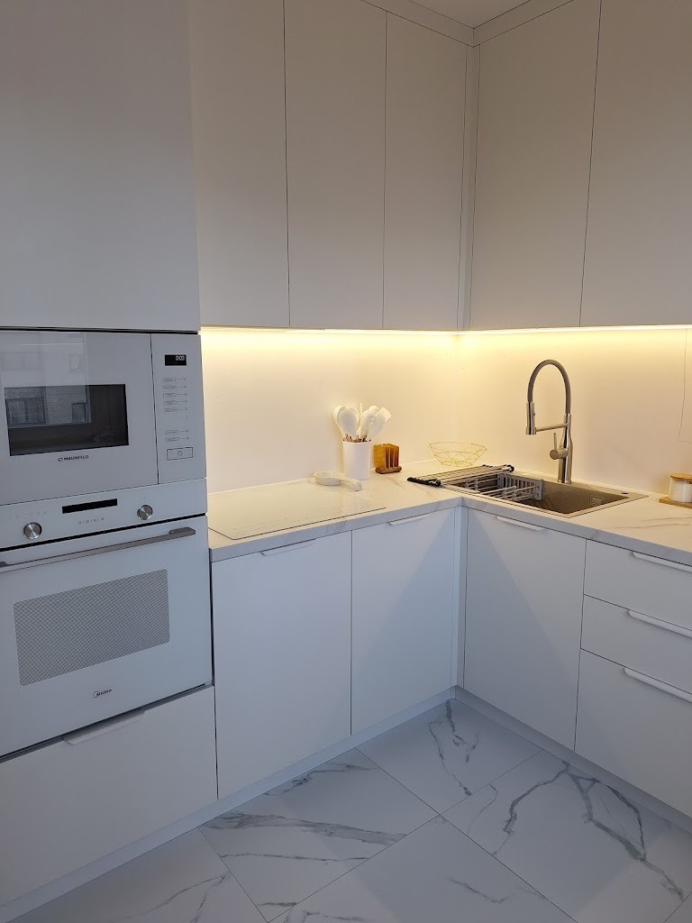 Minimalistyczna, biała kuchnia narożna z oświetleniem LED pod szafkami, marmurową podłogą i zabudowanym sprzętem AGD marki Midea i Maunfeld.
