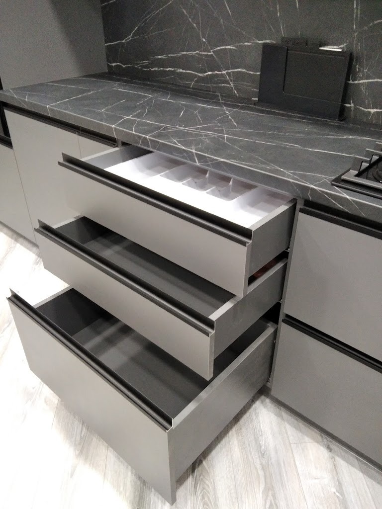 Szare, nowoczesne szafki kuchenne z otwartymi szufladami, widoczny wkład na sztućce w górnej szufladzie, blat z marmurowym wzorem.