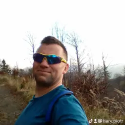 Mężczyzna w okularach przeciwsłonecznych, z plecakiem, na tle jesiennego krajobrazu z drzewami i wysoką trawą. Widoczny profil użytkownika na dole zdjęcia.