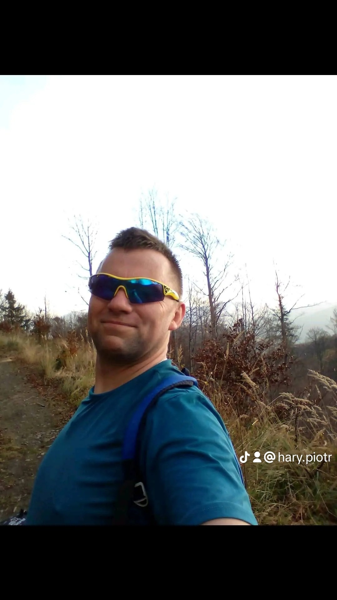 Mężczyzna w okularach przeciwsłonecznych, z plecakiem, na tle jesiennego krajobrazu z drzewami i wysoką trawą. Widoczny profil użytkownika na dole zdjęcia.